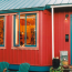 An exterior view of a small red cottage with blue metal roof and two adirondack chairs in front.